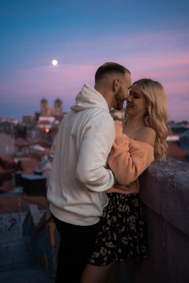 Tereza a Adam nad střechami Porta, nad nimi měsíc v růžovém soumraku